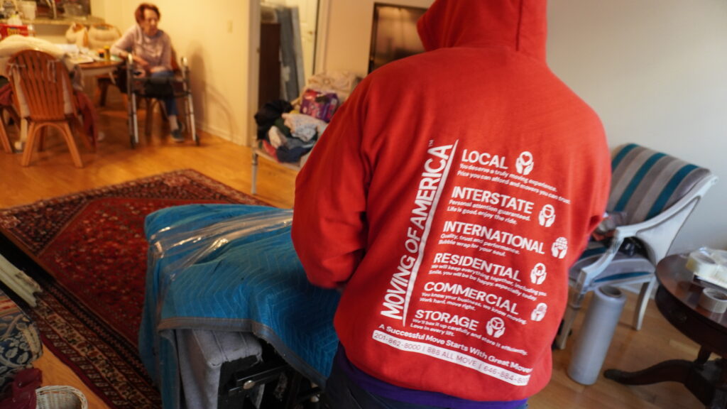 Residential mover in New Jersey protecting furniture with padding during a home move, with a senior resident in the background.